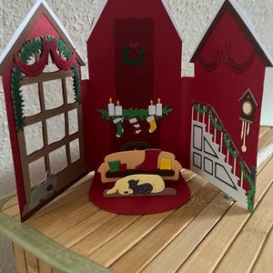 May include: A red Christmas-themed papercraft house with a central fireplace, a sofa with a sleeping dog, and side panels depicting a window and stairs. The house is decorated with garlands, candles, and a clock.