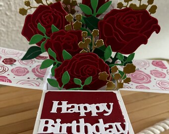 Könnte beinhalten: Eine rote und goldene Pop-up-Karte mit einem Strauß roter Rosen und grünen Blättern. Die Karte hat einen weißen Hintergrund mit einem rosa-weißen Muster und steht auf der Vorderseite "Happy Birthday".
