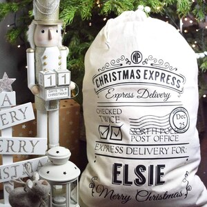 以下が含まれることがあります： 白いキャンバス地のクリスマス袋に、黒い文字で「Christmas Express Express Delivery Checked Twice North Pole Post Office Express Delivery For: Elsie Merry Christmas」と書いてあります。袋には紐で閉じる留め具と、「Dec 25」と書かれた赤と白のスタンプが付いています。