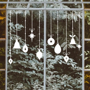 May include: White silhouette Christmas ornaments hanging in a window with a view of green foliage.