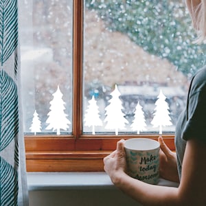 May include: A person holding a white mug with the text "Make today awesome" on it, looking out a window with a snowy scene. The window has white paper cutouts of pine trees on it.