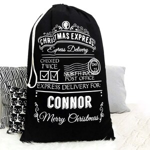 Op de afbeelding: Zwarte kerstcadeauzak met witte tekst. De tekst luidt "CHRISTMAS EXPRESS Express Delivery" en "CONNOR Merry Christmas". Voorzien van een trekkoord en feestelijke afbeeldingen.