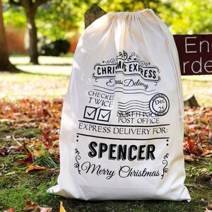 Op de afbeelding: Een cr&egrave;mekleurige kerstcadeauzak met zwarte letters. De tekst luidt "Christmas Express" en "Express Delivery For: Spencer Merry Christmas." De zak is vastgebonden met een trekkoord en ligt op gras en gevallen bladeren.