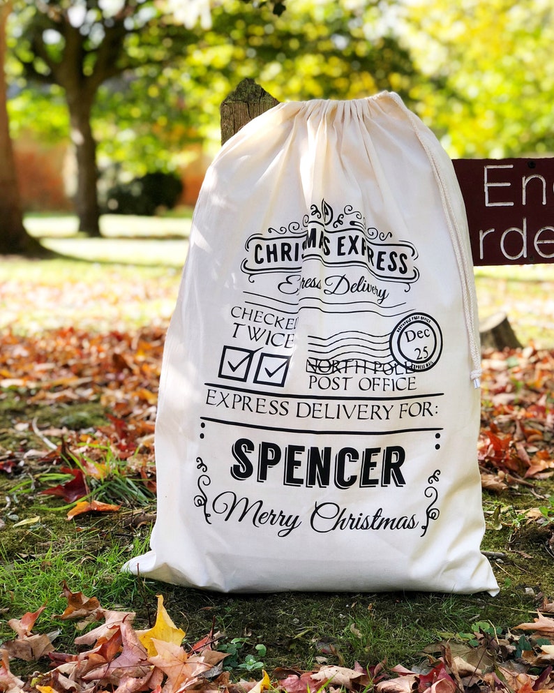 Op de afbeelding: Een cr&egrave;mekleurige kerstcadeauzak met zwarte letters. De tekst luidt "Christmas Express" en "Express Delivery For: Spencer Merry Christmas." De zak is vastgebonden met een trekkoord en ligt op gras en gevallen bladeren.