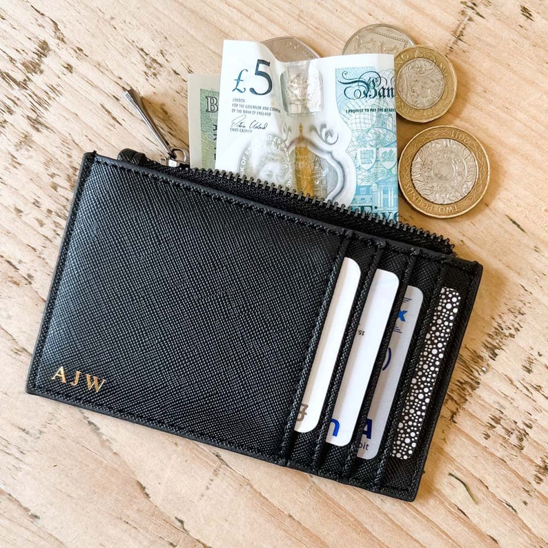 K&ouml;nnte beinhalten: Eine schwarze Ledergeldb&ouml;rse mit Rei&szlig;verschluss und einem goldenen, gepr&auml;gten Monogramm "AJW". Die Geldb&ouml;rse hat mehrere Kartenf&auml;cher und ist ge&ouml;ffnet, um Bargeld und Karten im Inneren zu zeigen.