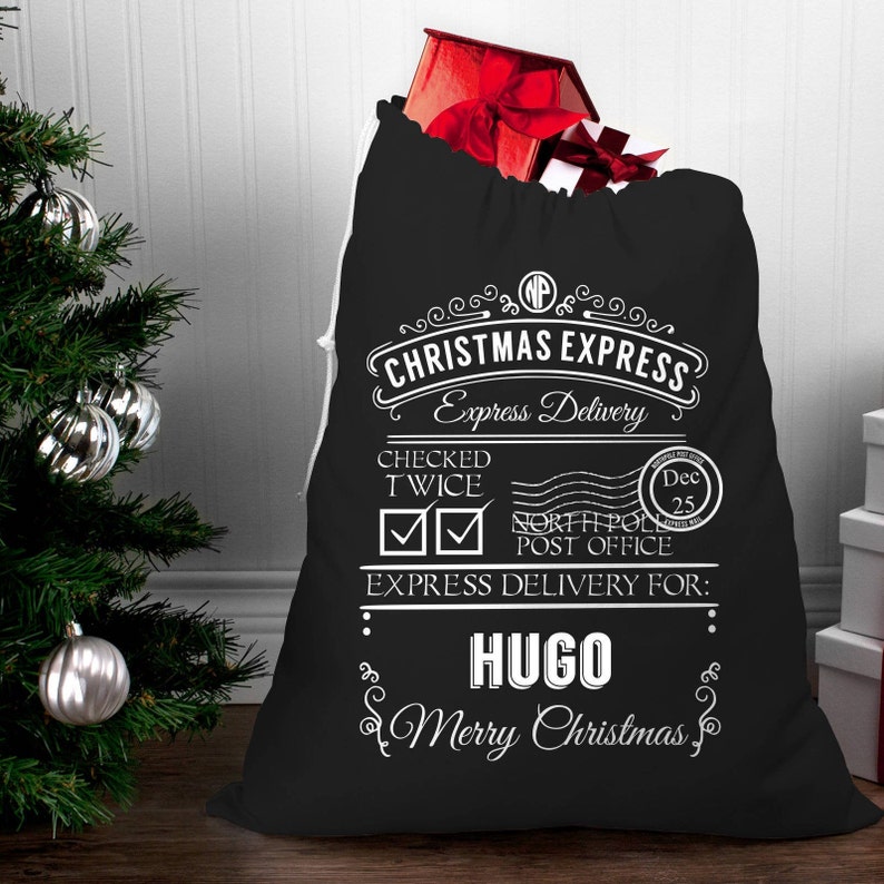 Op de afbeelding: Zwarte kerstzak met wit trekkoord, gevuld met een rode geschenkdoos. De zak heeft de tekst "CHRISTMAS EXPRESS" en "EXPRESS DELIVERY FOR: HUGO, Merry Christmas". Een kerstboom met zilveren ballen staat op de achtergrond.