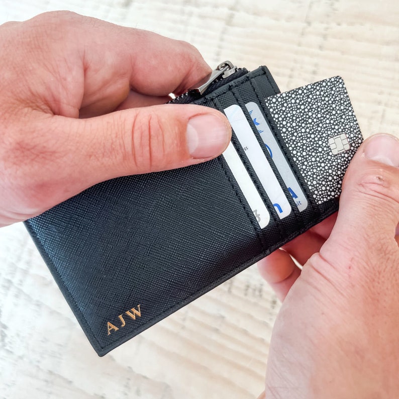 K&ouml;nnte beinhalten: Eine schwarze Ledergeldb&ouml;rse mit Rei&szlig;verschluss und den in Gold gepr&auml;gten Initialen "AJW". Die Geldb&ouml;rse ist ge&ouml;ffnet und zeigt drei Karten im Inneren.