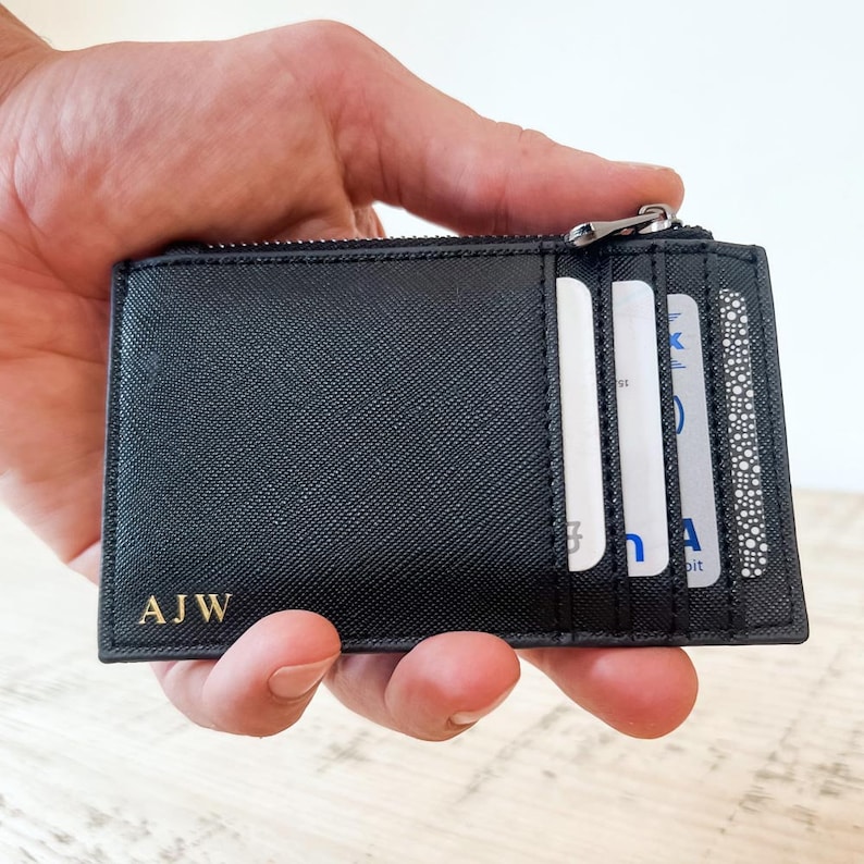K&ouml;nnte beinhalten: Schwarze Ledergeldb&ouml;rse mit Rei&szlig;verschluss und einem goldenen gepr&auml;gten Monogramm "AJW". Die Geldb&ouml;rse hat einen Rei&szlig;verschluss und mehrere Kartenf&auml;cher.