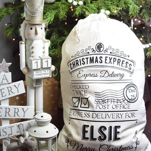Op de afbeelding: Een kersttafereel met een cr&egrave;mekleurige zak met de tekst "Christmas Express" en "Elsie". Een houten notenkrakerfiguur en een decoratief bord met de tekst "Merry Christmas" zijn ook aanwezig, met een kerstboom op de achtergrond.