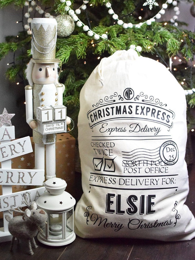 Op de afbeelding: Een kersttafereel met een cr&egrave;mekleurige zak met de tekst "Christmas Express" en "Elsie". Een houten notenkrakerfiguur en een decoratief bord met de tekst "Merry Christmas" zijn ook aanwezig, met een kerstboom op de achtergrond.