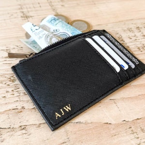 K&ouml;nnte beinhalten: Ein schwarzer Lederkartenhalter mit Rei&szlig;verschluss und goldenen Initialen "AJW" auf der Vorderseite. Der Kartenhalter ist ge&ouml;ffnet und enth&auml;lt Bargeld und Kreditkarten.