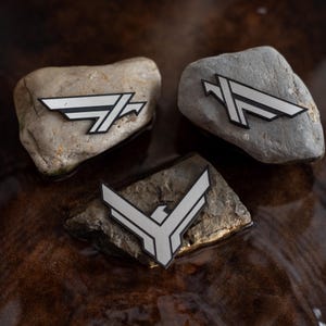 May include: Three silver and black emblem patches with a stylized bird design are displayed on natural stones. The patches have a geometric aesthetic and are set against a dark, textured background, creating a contrast between the smooth patches and the rough stones.