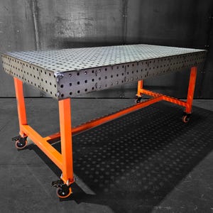 May include: A metal welding table with a perforated top and an orange metal frame. The table has four wheels and is on a gray concrete floor.