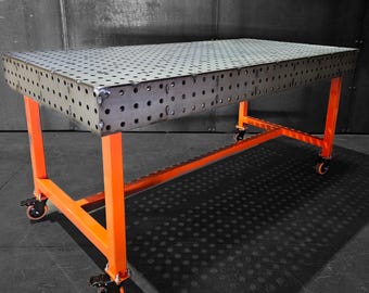May include: A metal welding table with a perforated top and an orange metal frame. The table has four wheels and is on a gray concrete floor.