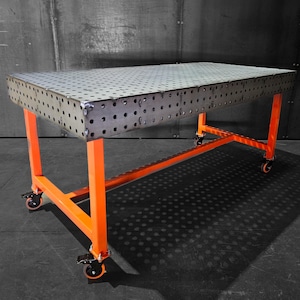 May include: A metal welding table with a perforated top and orange painted legs. The table is on wheels and has a metal frame.