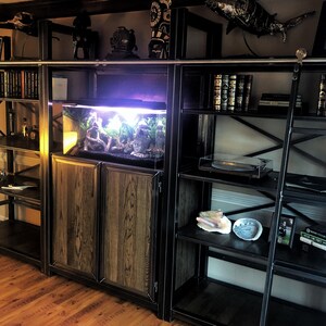 May include: A black metal shelving unit with a wooden cabinet and a fish tank. The shelves are filled with books, records, and decorative items. The cabinet has two doors and is made of wood with a dark stain. The fish tank is lit and has a variety of plants and rocks inside.