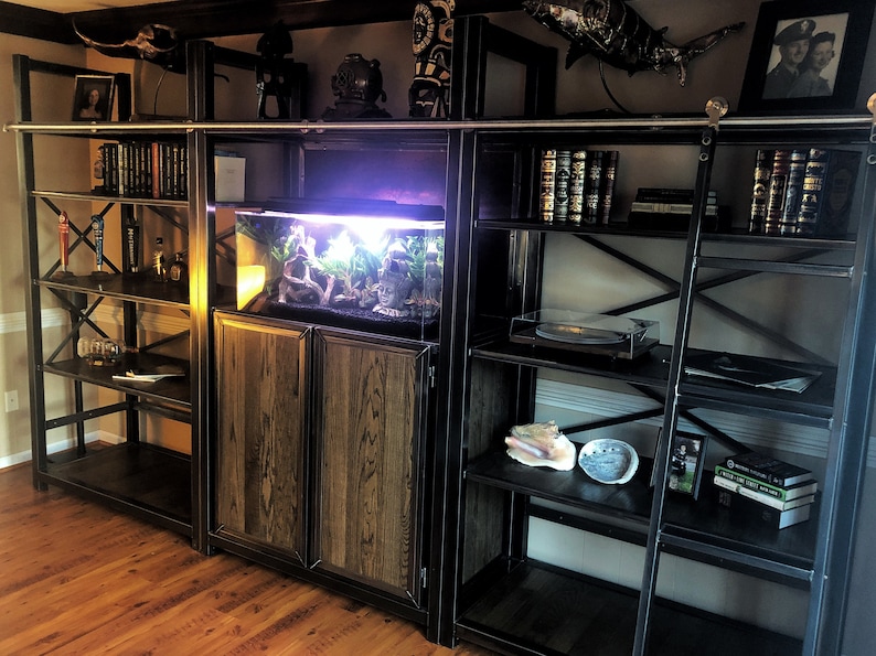 May include: A black metal shelving unit with a wooden cabinet and a fish tank. The shelves are filled with books, records, and decorative items. The cabinet has two doors and is made of wood with a dark stain. The fish tank is lit and has a variety of plants and rocks inside.