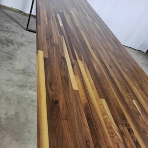 May include: A close-up view of a wooden table top made of walnut planks. The wood has a rich brown color with a natural grain pattern. The table top is smooth and polished.