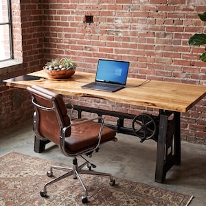 Puede incluir: Un escritorio de madera con borde natural en acabado marrón claro y una base industrial de metal negro con un mecanismo de ajuste de manivela. Un portátil gris oscuro se encuentra sobre la superficie junto a un cuenco de cobre con plantas.