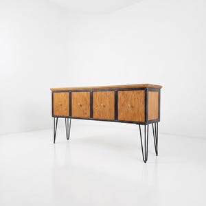 May include: A rectangular wooden sideboard featuring four cabinet doors with light brown wood grain, a black metal frame, and four black metal hairpin legs. Each cabinet door is equipped with a small, round, metallic knob for opening.