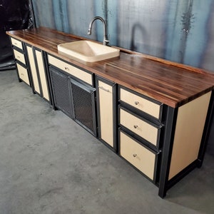 Puede incluir: Una isla de cocina moderna con una encimera de nogal, un marco de metal negro y un fregadero de porcelana blanca. La isla cuenta con un armario con una puerta de malla met&aacute;lica, cajones y un armario con una puerta s&oacute;lida.
