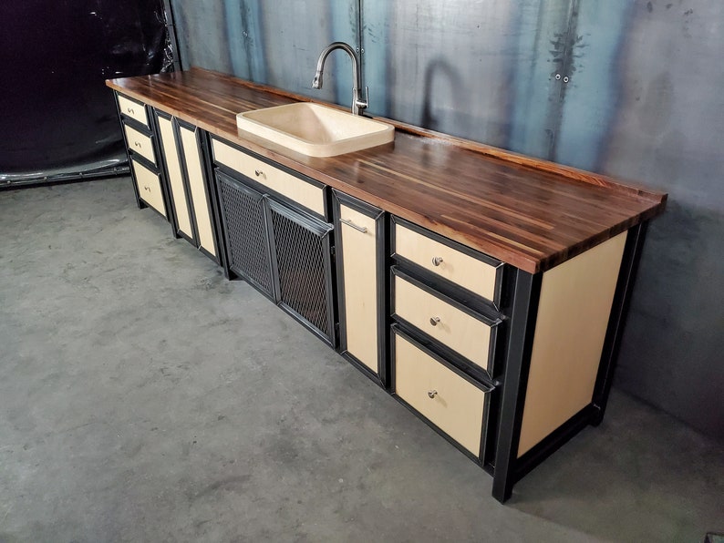 Puede incluir: Una isla de cocina moderna con una encimera de nogal, un marco de metal negro y un fregadero de porcelana blanca. La isla cuenta con un armario con una puerta de malla met&aacute;lica, cajones y un armario con una puerta s&oacute;lida.