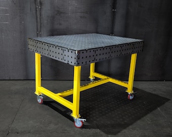 May include: A black metal welding table with a perforated top and yellow metal legs. The table is on wheels and has a gray background.