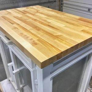 Peut inclure: Un meuble blanc avec un plan de travail en bois. Le meuble a deux portes avec des panneaux en verre et des poignées en métal. Le plan de travail est en bois clair.