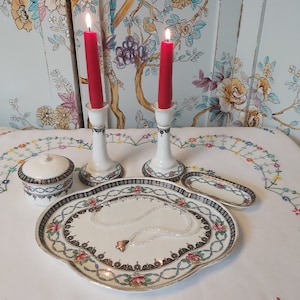 Puede incluir: Un juego de tocador de porcelana blanca con un diseño floral decorativo. El juego incluye dos candelabros con velas rojas, un plato pequeño redondo, un plato rectangular y un plato grande ovalado con un collar de perlas.