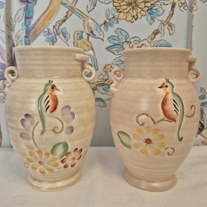 Peut inclure: Une paire de vases en céramique de couleur crème avec des corps nervurés et de petites anses latérales. Chaque vase présente un motif peint à la main d'un petit oiseau perché sur une branche avec des fleurs jaunes, violettes et rouges.