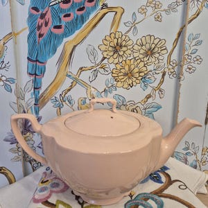 Può includere: Una teiera in ceramica rosa chiaro con coperchio, beccuccio e manico abbinati. È posizionata su un panno bianco ricamato con colorati disegni floreali. Lo sfondo presenta una parete dipinta con un motivo di uccelli e fiori.