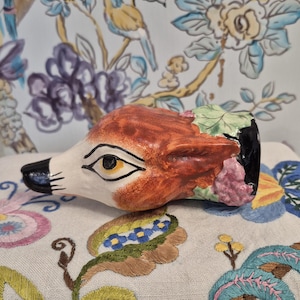 Puede incluir: Una figura de cerámica con forma de cabeza de zorro, pintada a mano con detalles en naranja, blanco y negro. El zorro tiene una cara detallada con un ojo amarillo y está decorado con detalles en rosa y verde. La figura descansa sobre una superficie bordada floral.