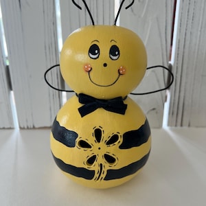 May include: A handcrafted, whimsical bee figurine. The bee is painted yellow with black stripes and a black bow tie. It has a smiling face, antennae, and wings. The lower body features a decorative flower cut-out.