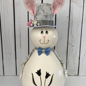 May include: A decorative Easter bunny figurine with a cream-colored body, a top hat, and a blue bow tie. The bunny has large, upright ears with pink accents and a smiling face. The body features a tulip design.