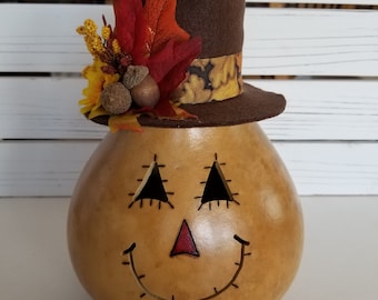 May include: A brown gourd with a painted face and a stitched smile. The gourd is wearing a brown top hat with a yellow and brown patterned band and a fall-themed floral arrangement.