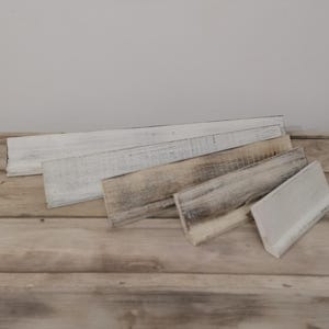 May include: Five wooden signs of varying sizes, all painted white with a distressed finish. The signs are arranged in a staggered pattern on a wooden surface.