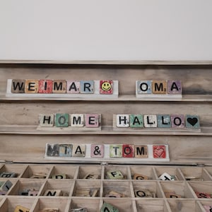 May include: A wooden box with a hinged lid, containing a collection of letter tiles. The tiles are arranged in compartments and feature various letters and numbers. The box is open, revealing the tiles inside. The tiles are painted in various colors and have a distressed, vintage look. The box is labeled with the words "WEIMAR", "OMA", "HOME", "HALLO", "MIA", and "TOM".