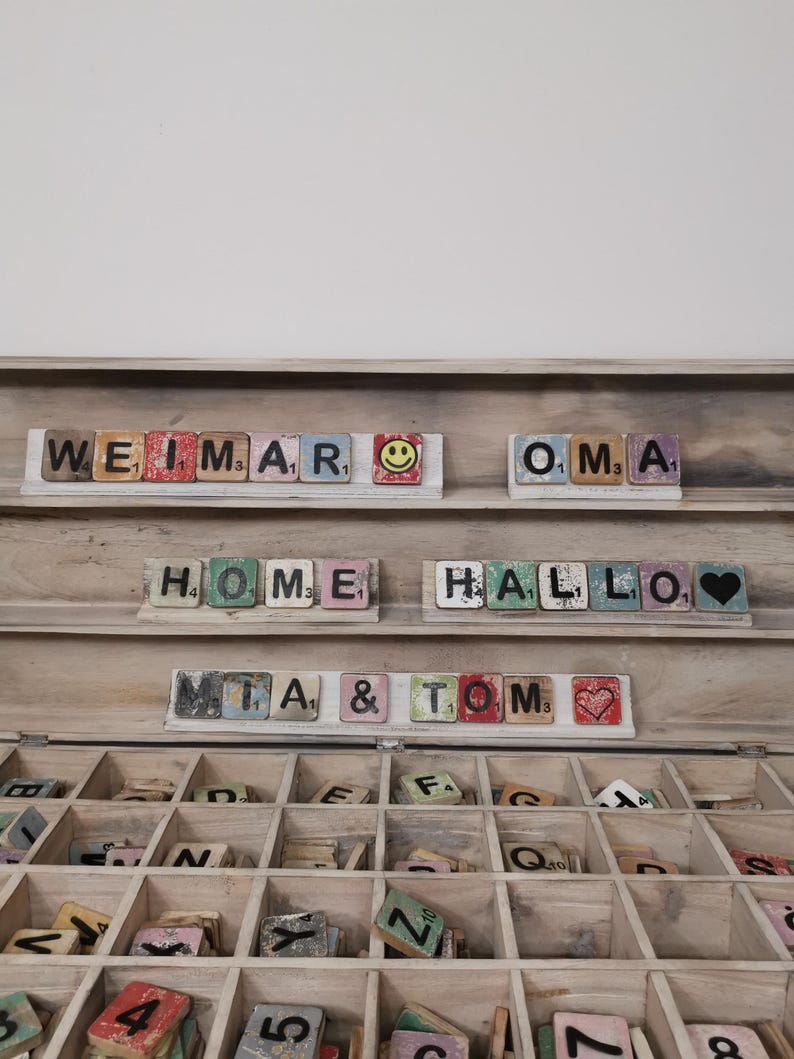 May include: A wooden box with a hinged lid, containing a collection of letter tiles. The tiles are arranged in compartments and feature various letters and numbers. The box is open, revealing the tiles inside. The tiles are painted in various colors and have a distressed, vintage look. The box is labeled with the words "WEIMAR", "OMA", "HOME", "HALLO", "MIA", and "TOM".