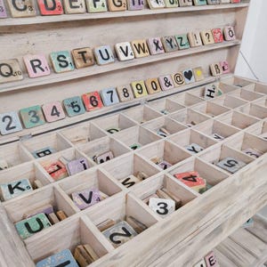 May include: A wooden box with a whitewashed finish, containing a collection of vintage letter tiles. The tiles are arranged in alphabetical order and include numbers, punctuation marks, and symbols. The box has a two-tiered design, with the top tier containing the letters and the bottom tier containing the numbers and symbols.