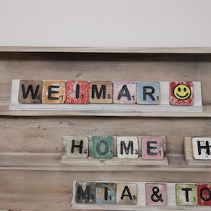 May include: A wooden box with a shelf and compartments. The shelf has a row of colorful wooden tiles spelling out "WEIMAR" with a smiley face tile at the end. The next shelf has tiles spelling out "HOME" and "H". The bottom shelf has tiles spelling out "MIA & TO".
