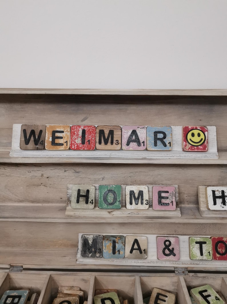 May include: A wooden box with a shelf and compartments. The shelf has a row of colorful wooden tiles spelling out "WEIMAR" with a smiley face tile at the end. The next shelf has tiles spelling out "HOME" and "H". The bottom shelf has tiles spelling out "MIA & TO".