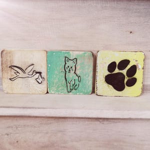 May include: Three wooden blocks with different images. The first block is white with a black stork carrying a baby in its beak. The second block is green with a black cat. The third block is yellow with a black paw print.