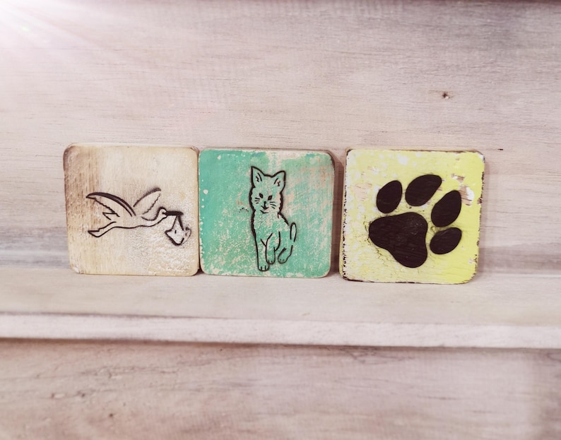 May include: Three wooden blocks with different images. The first block is white with a black stork carrying a baby in its beak. The second block is green with a black cat. The third block is yellow with a black paw print.