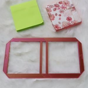 Könnte beinhalten: Ein rosa Plastikrahmen mit zwei rechteckigen Öffnungen. Der Rahmen liegt auf einer weißen, flauschigen Oberfläche. Im Hintergrund befinden sich ein grüner Haftnotiz und eine Blumen-Geschenkbox.