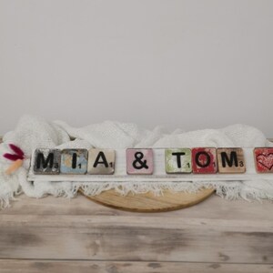 May include: A white wooden sign with the names "MIA & TOM" spelled out in black letters on colorful tiles. A red heart tile is at the end of the sign.