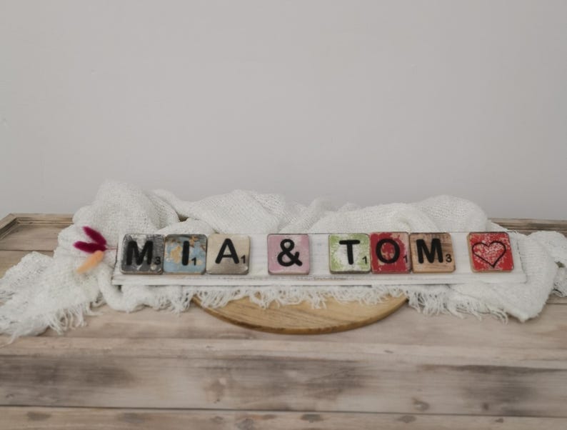 May include: A white wooden sign with the names "MIA & TOM" spelled out in black letters on colorful tiles. A red heart tile is at the end of the sign.