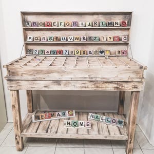 May include: A wooden table with a shelf and a compartment for storing wooden letter blocks. The blocks are arranged in alphabetical order and include numbers, punctuation marks, and symbols. The table is painted white and has a distressed finish. The words "WEIMAR", "HALLO", and "HOME" are spelled out on the bottom shelf using the letter blocks.