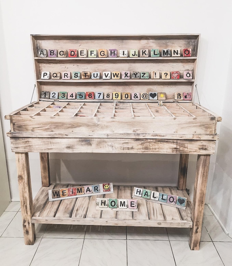 May include: A wooden table with a shelf and a compartment for storing wooden letter blocks. The blocks are arranged in alphabetical order and include numbers, punctuation marks, and symbols. The table is painted white and has a distressed finish. The words "WEIMAR", "HALLO", and "HOME" are spelled out on the bottom shelf using the letter blocks.