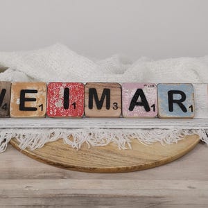 May include: A wooden sign spelling out the word "WEIMAR" using individual letter tiles. Each tile is painted in a different color and has a number on it. The sign is sitting on a white surface with a white blanket draped over it.