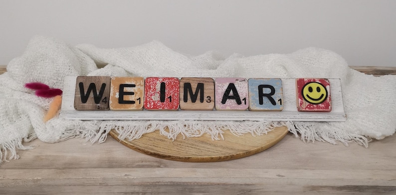 May include: A wooden sign spelling out the word "WEIMAR" using individual letter tiles. Each tile is painted in a different color and has a number on it. The sign is sitting on a white surface with a white blanket draped over it.